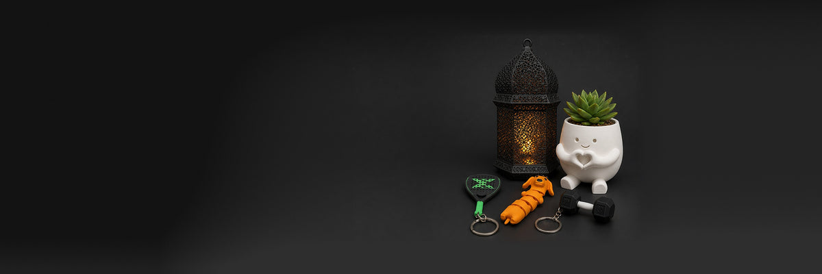 Regalos impresos en 3D de IVARO sobre una mesa oscura: farol árabe encendido, maceta corazón sonriente con suculenta y llaveros de pala de pádel, perro salchicha y mancuerna