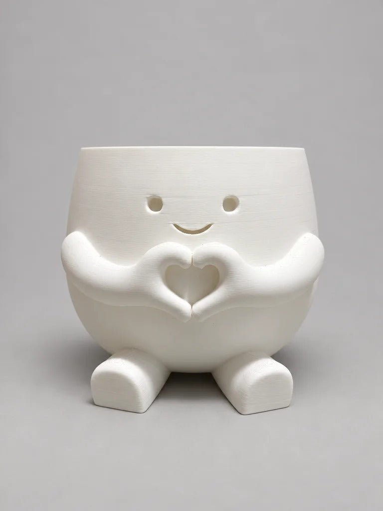 Maceta blanca con cara sonriente y dos manitas formando un corazón, apoyada sobre dos patitas