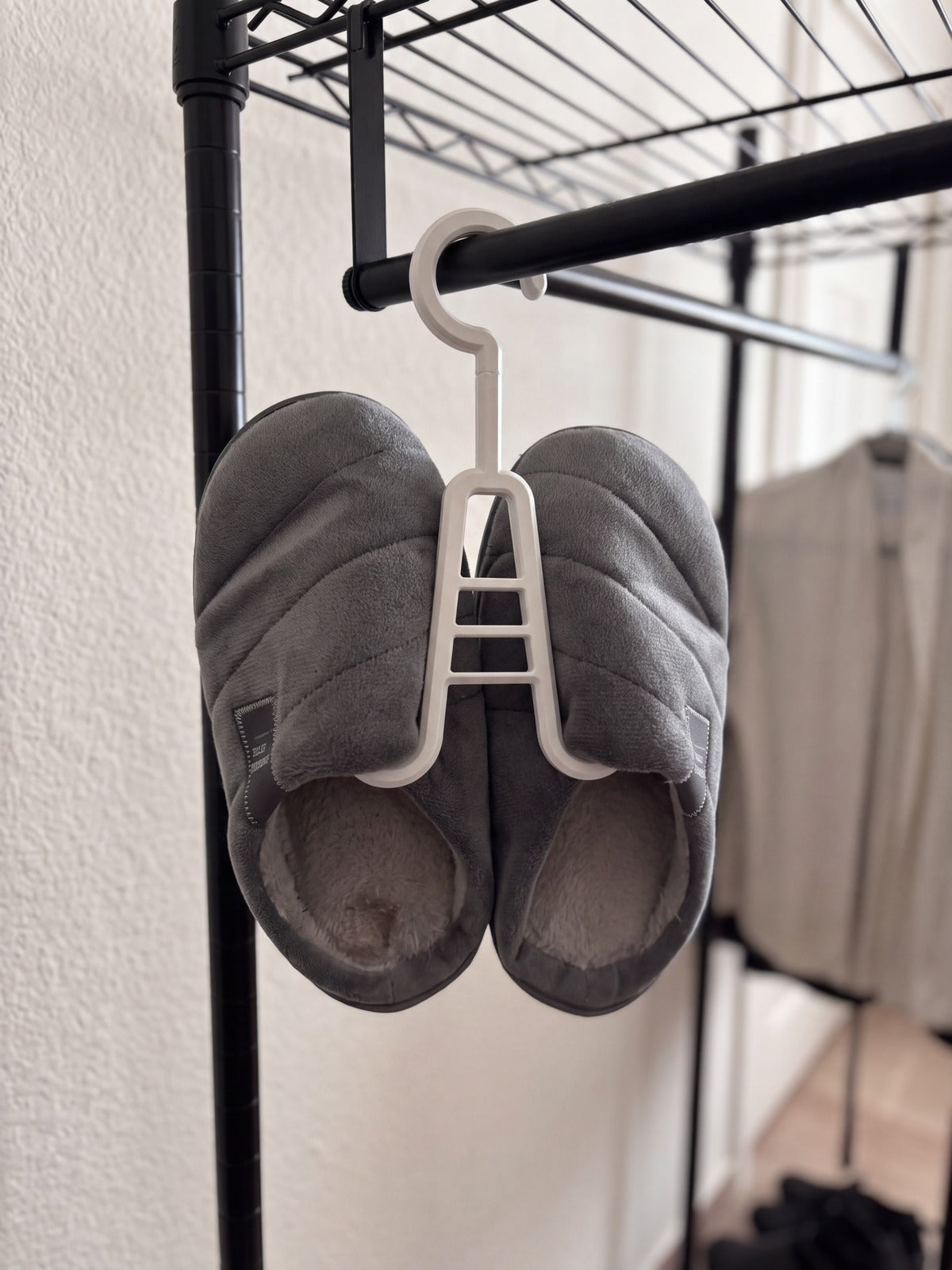 Percha blanca colgada de la barra de un armario sujetando por dentro un par de zapatillas de estar por casa grises (imagen generada con IA)