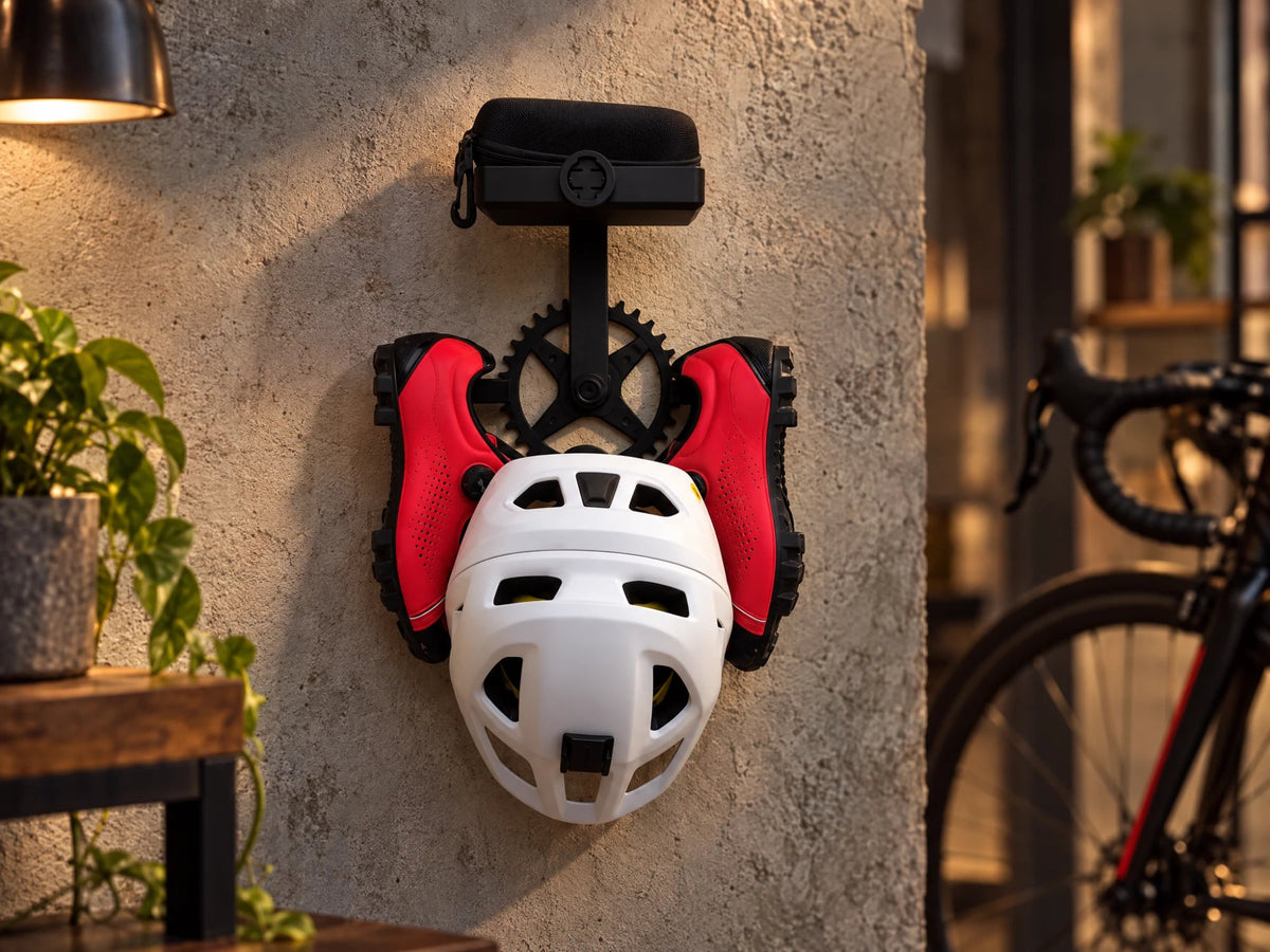 Soporte de pared negro con forma de plato y biela de bicicleta sujetando un casco, unas zapatillas de ciclismo y una funda